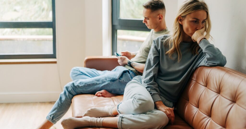 A woman on a couch looks down and has her back to the man on the cushion beside her. The man looks at his cell phone.