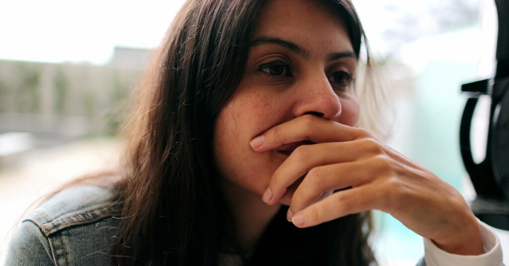 A woman holds her hand over her mouth with the fingers slightly apart. She looks ahead of her with unfocused eyes.