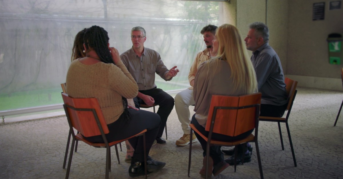 A small group of people sit in chairs at an al-anon meeting. One individual is actively speaking to the others.