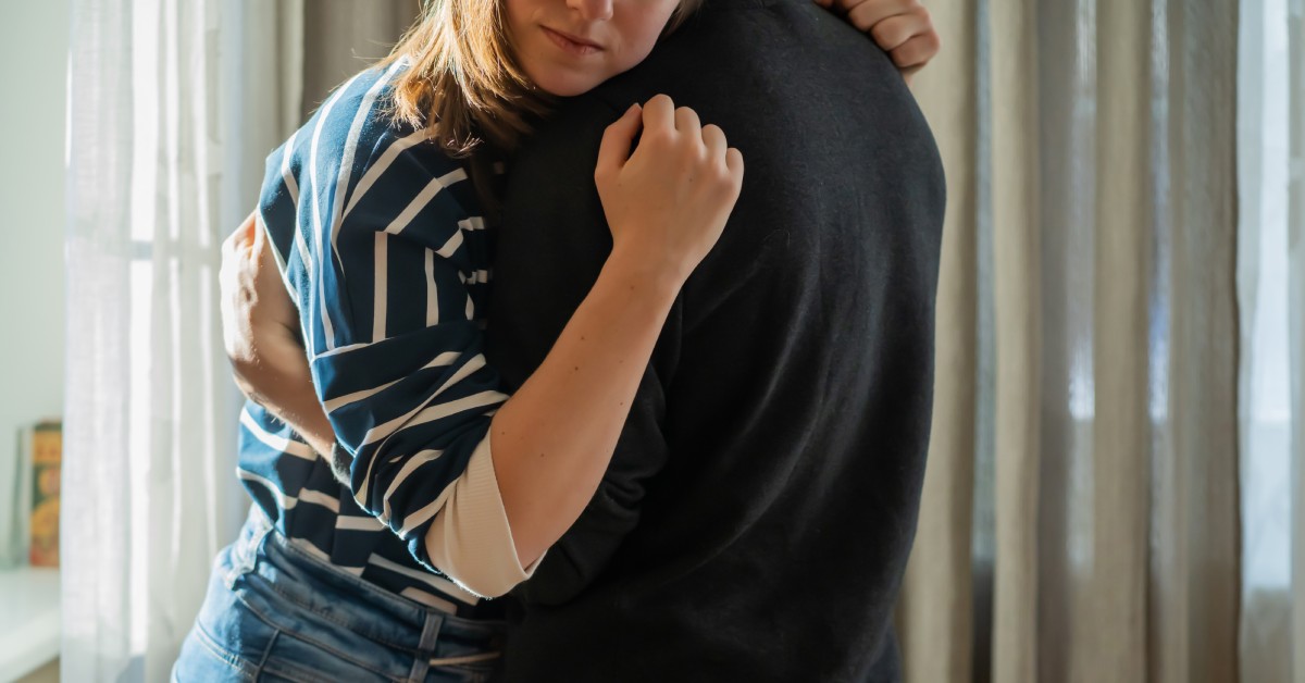 A pair of friends embrace one another in a somber hug. One wears a black top and the other wears a navy and white striped top.