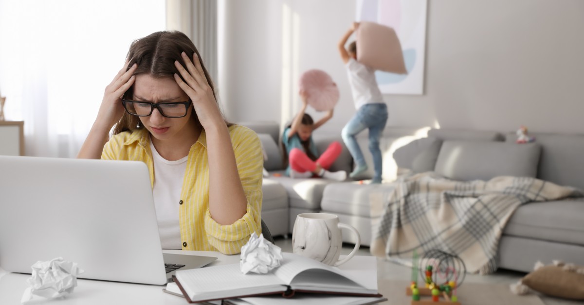 A stressed mom sits at the table in front of a laptop. Her two children fight on the couch behind her with pillows.