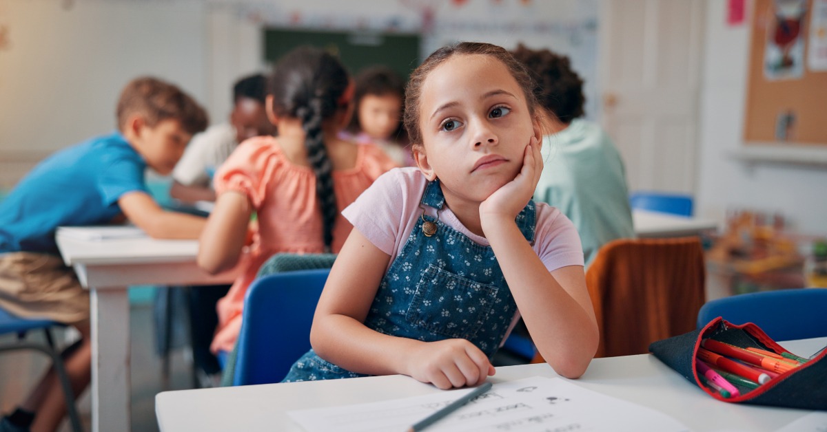 A young girl at a classroom desk rests her head on her hand and looks to the side. Other kids behind her work together.
