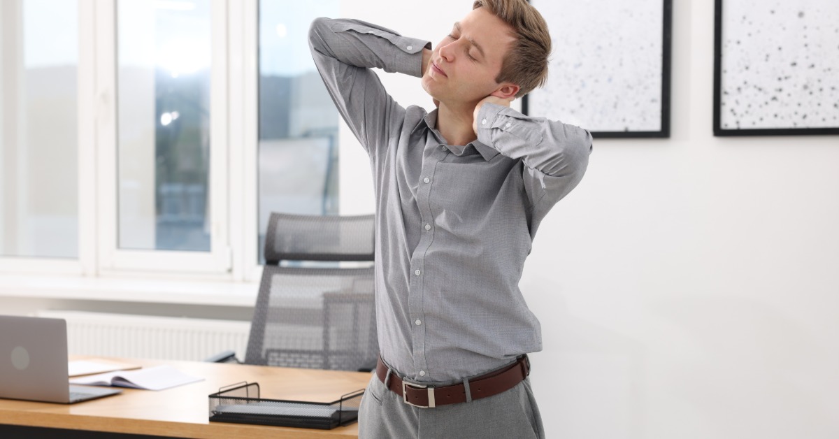 A man in a bright office presses both of his hands to the back of his neck as he stretches with his eyes closed.