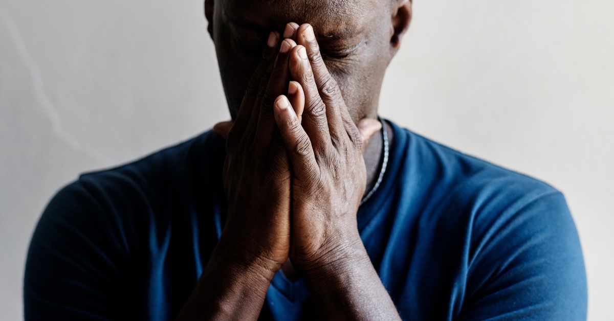 A man with closed eyes presses his palms together, covering his nose and mouth. He tilts his head slightly downward.