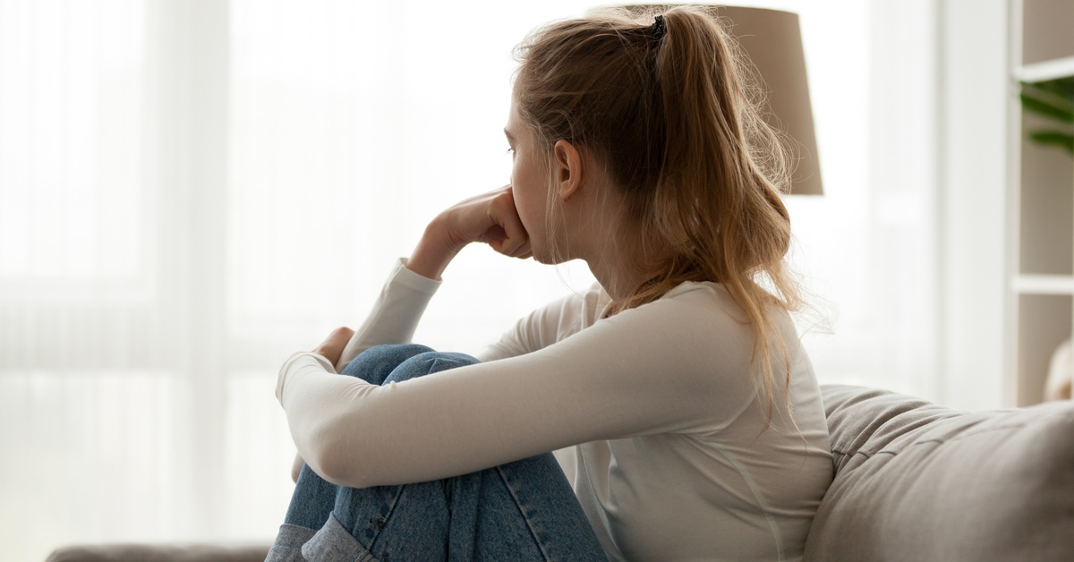 A woman sits on a couch with her knees pulled up. She rests her chin on her hand and looks toward a curtained window.