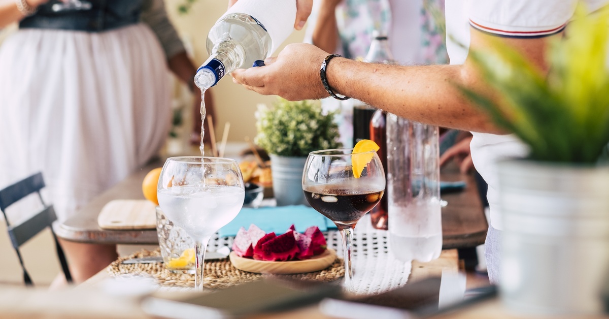 A person pours clear liquor from a bottle into a large stemmed glass that's on a table beside plates and another drink.