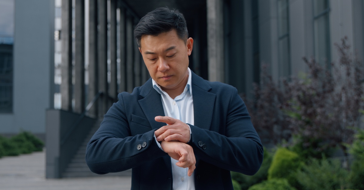 A man in business attire stands outside near a row of columns. He looks down at his right wrist and adjusts his watch.
