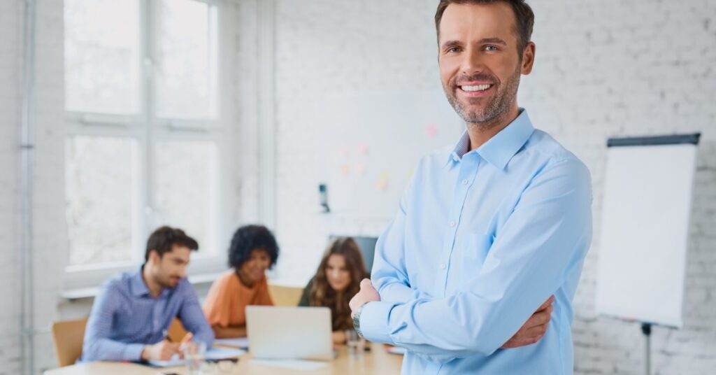 A smiling man stands with his arms crossed, looking at the viewer. Behind him, people sit at a table and work together.