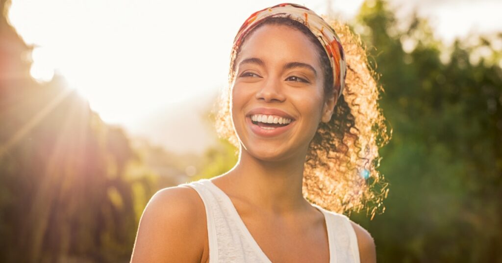 A woman standing in front of greenery smiles as she looks to the side. Sunlight shines from behind her curls.