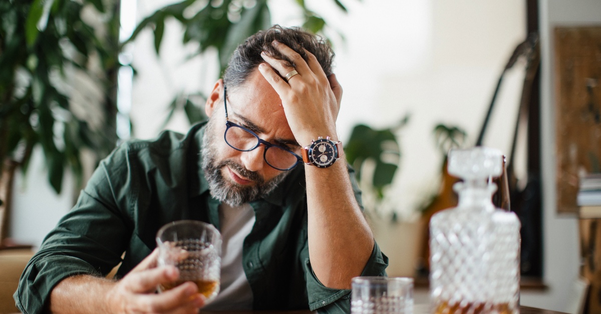 A man looks at a glass of amber liquid he holds. A decanter and another glass sit on the table in front of him.