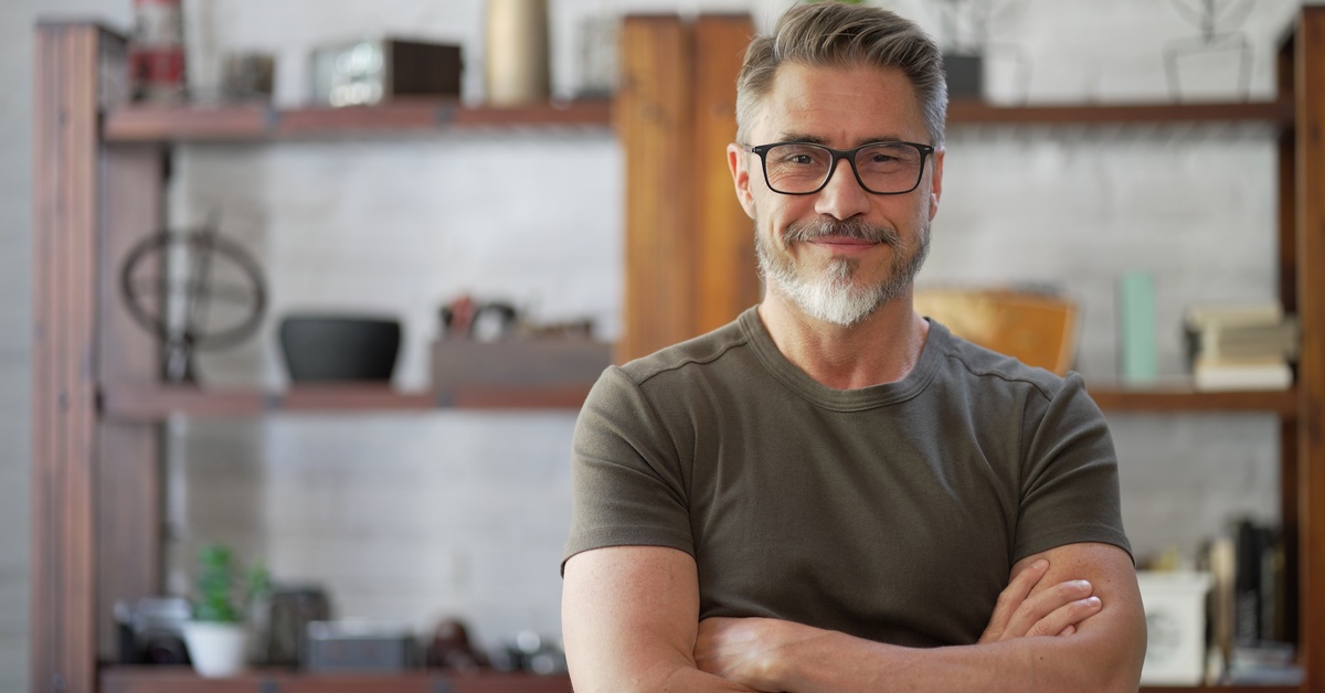 A smiling man with gray hair and glasses stands with his arms crossed. Shelves of decor are in the blurred background.