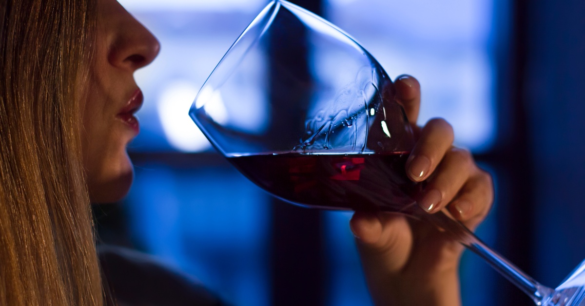 A person’s face is in profile as they raise a glass of red wine to their mouth. The background is blue and blurred.