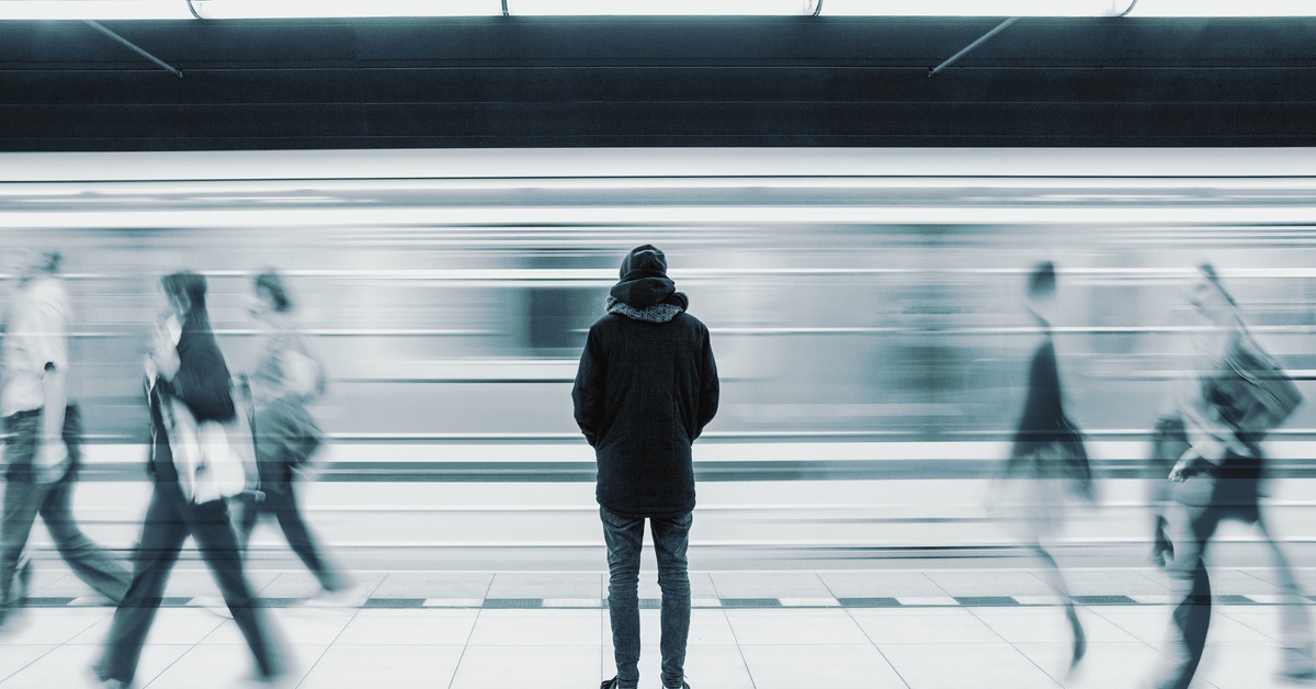 A person stands on a subway platform facing a passing train that appears blurred from motion. Blurred people walk past.