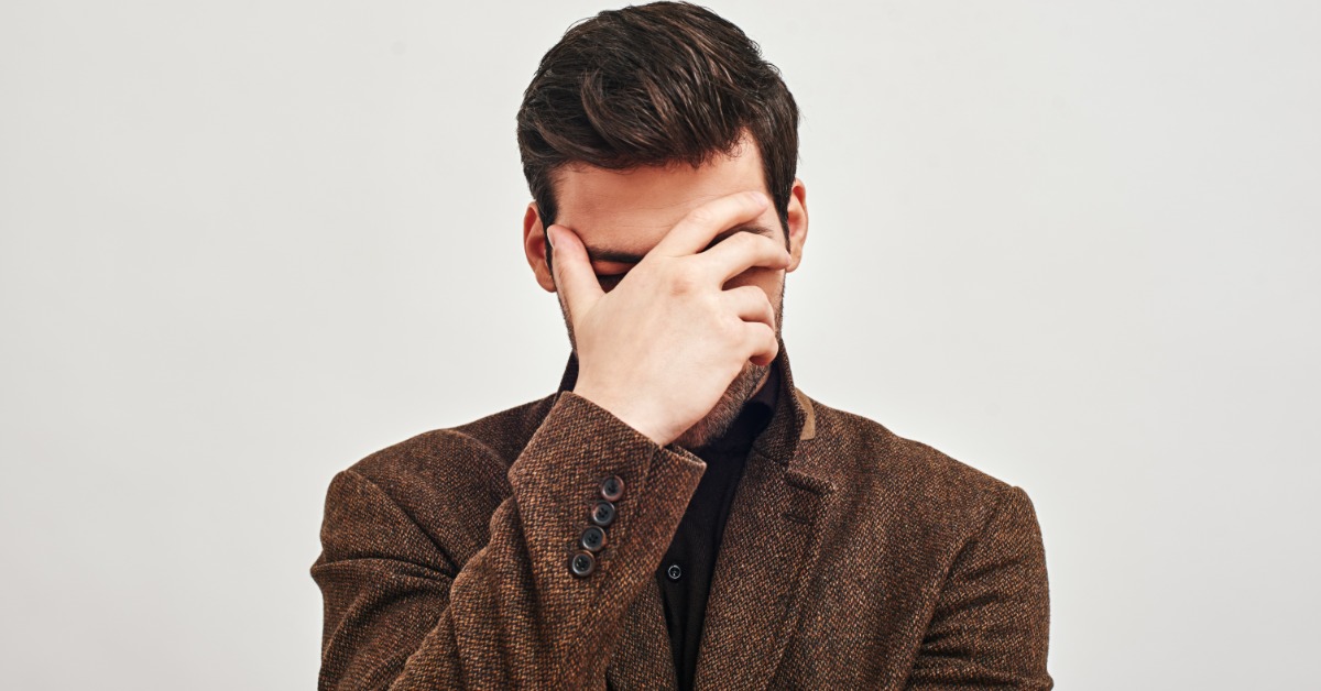 A man in a textured brown blazer stands against a plain background. His hand covers his face, which is tilted downward.