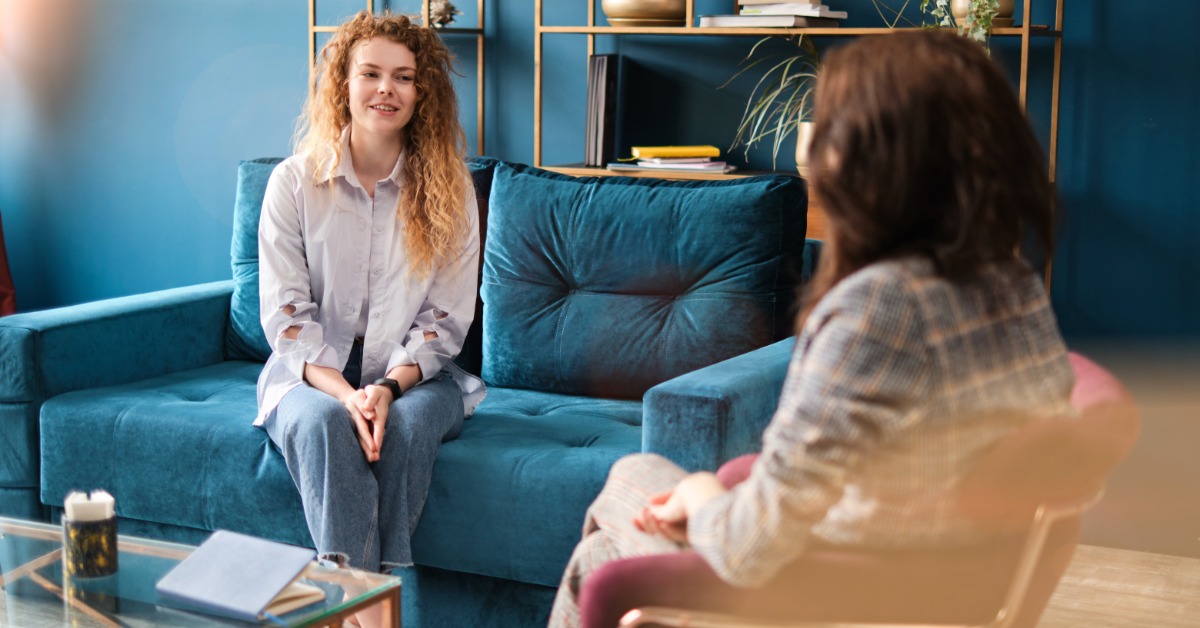 A woman on a couch faces another person seated in a chair. A closed notebook is on the coffee table between them.