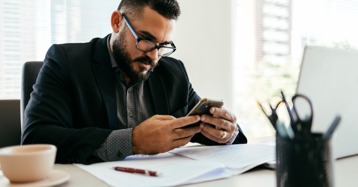 A man in a dark blazer sits at a desk and looks at his phone. Papers, a laptop, a pen, and a coffee cup are on the desk.