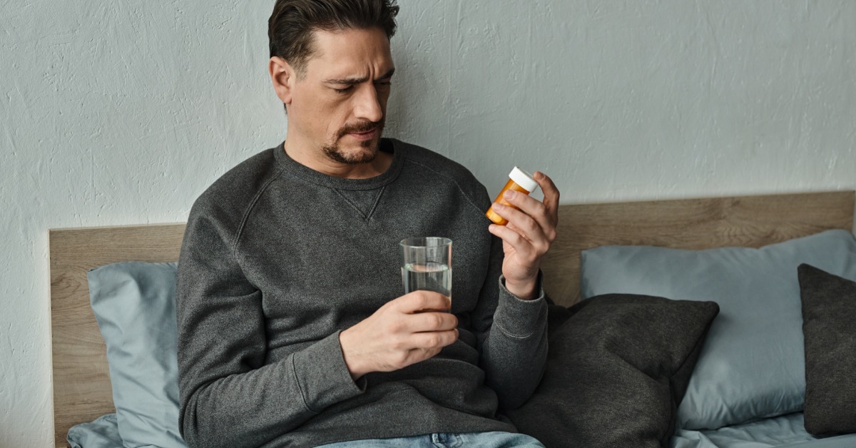 A man sits against a headboard, holding a prescription bottle and a glass of water. He frowns and looks at the bottle.