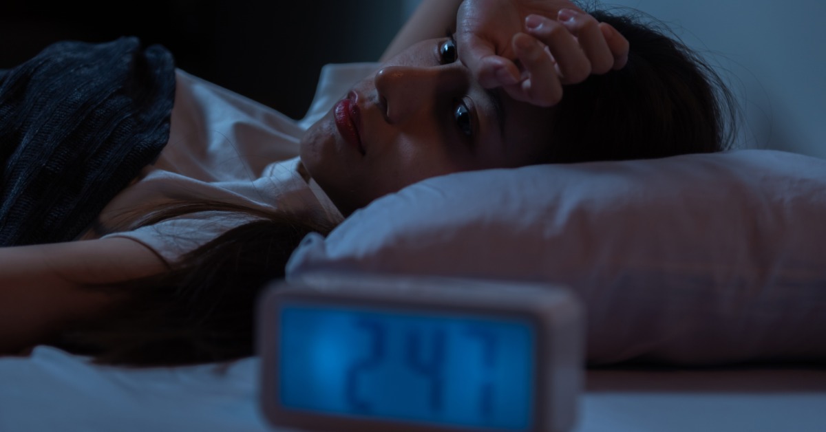 A woman lies in bed in low light with the back of one hand on her forehead. A blurred digital clock nearby reads 2:47.