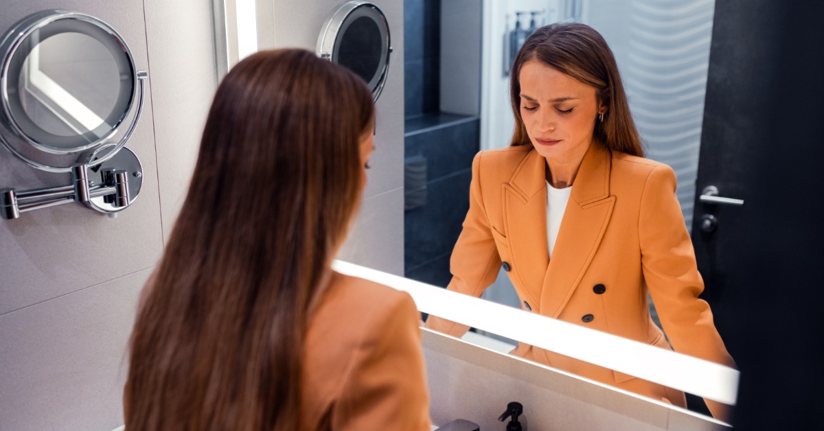A woman in a tan blazer stands at a bathroom sink, facing a large, illuminated mirror. Her eyes are closed.