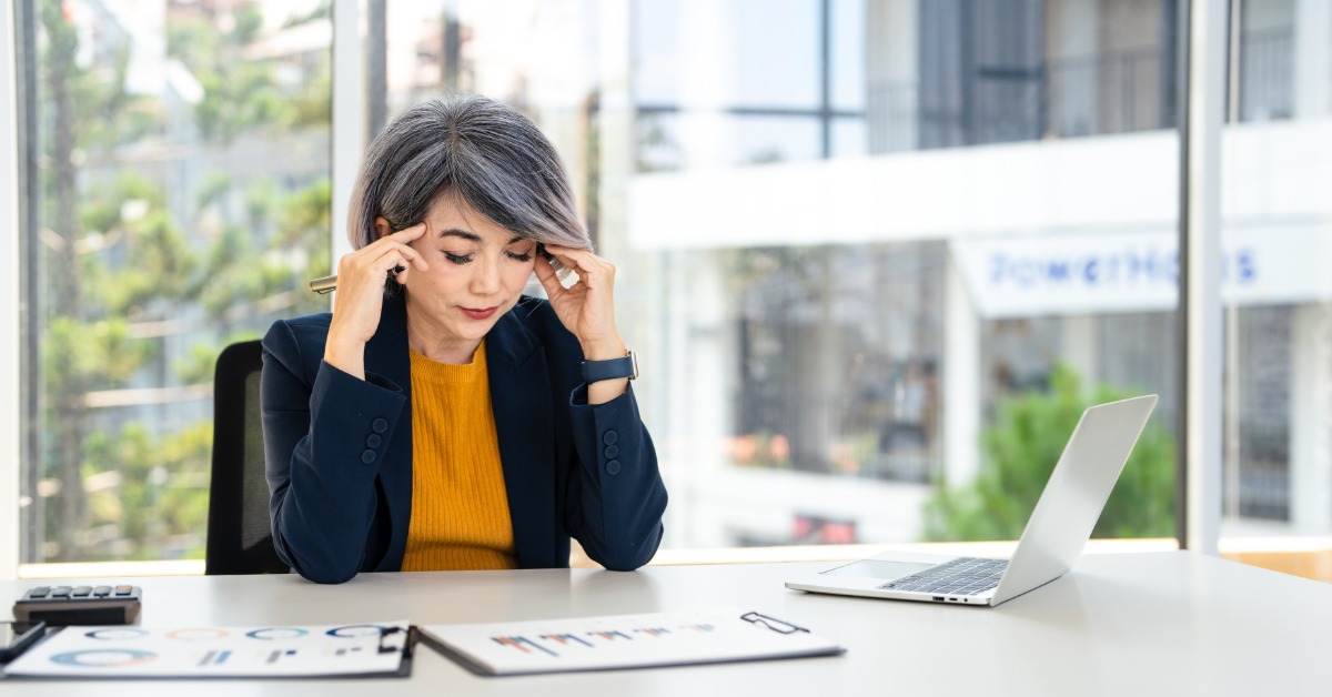 A woman presses her fingers to her temples. An open laptop and several printed charts are on the desk in front of her.