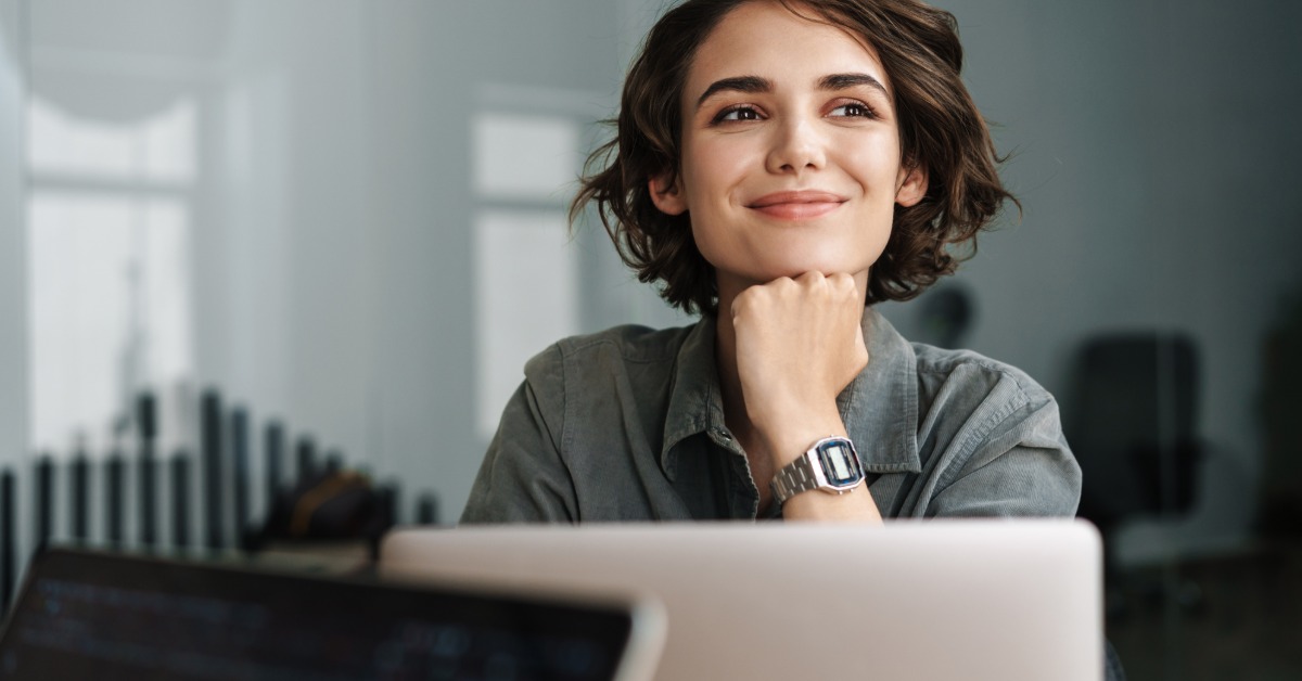 A smiling woman with short, dark hair sits at a desk, her chin resting on her hand. A laptop is open in front of her.