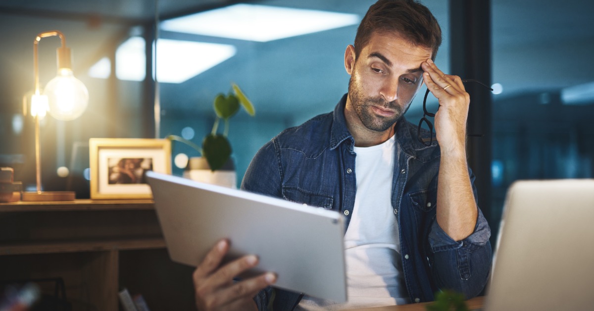 A man in an office looks at a tablet in his hand. With his other hand, he holds his glasses and touches his forehead.