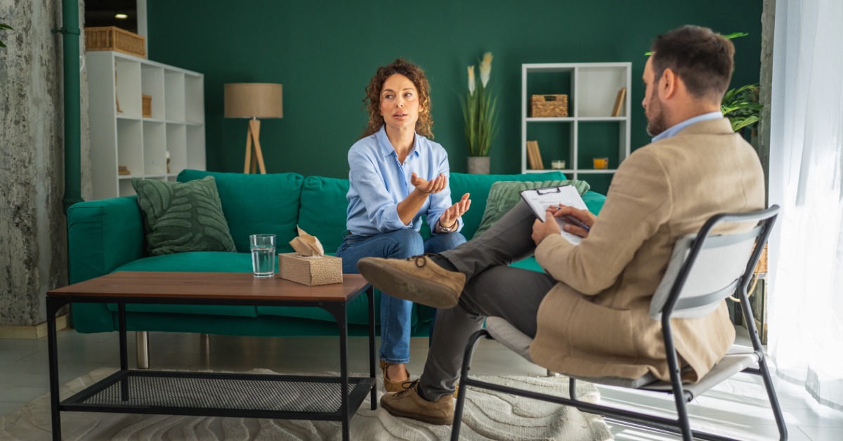 A woman leans forward on a green couch and gestures as she speaks to a man seated across from her. He holds a clipboard.