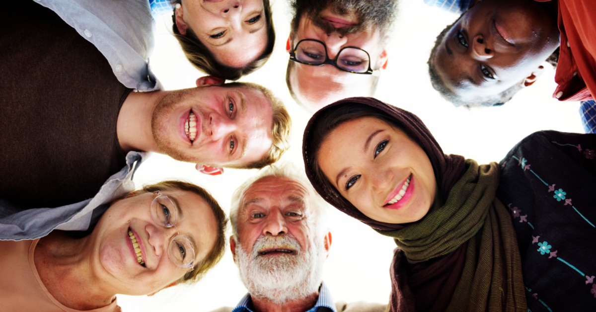 Upward view of seven adults standing in a circle and looking down. They are of different ages, races, and genders.