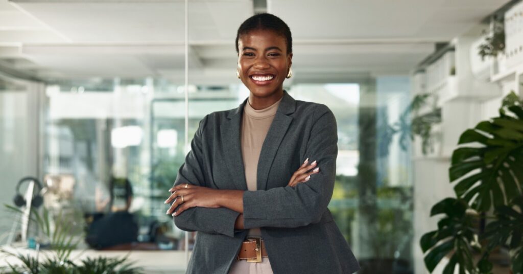 A smiling woman in a gray blazer stands with crossed arms in an office. Glass walls and plants are behind her.