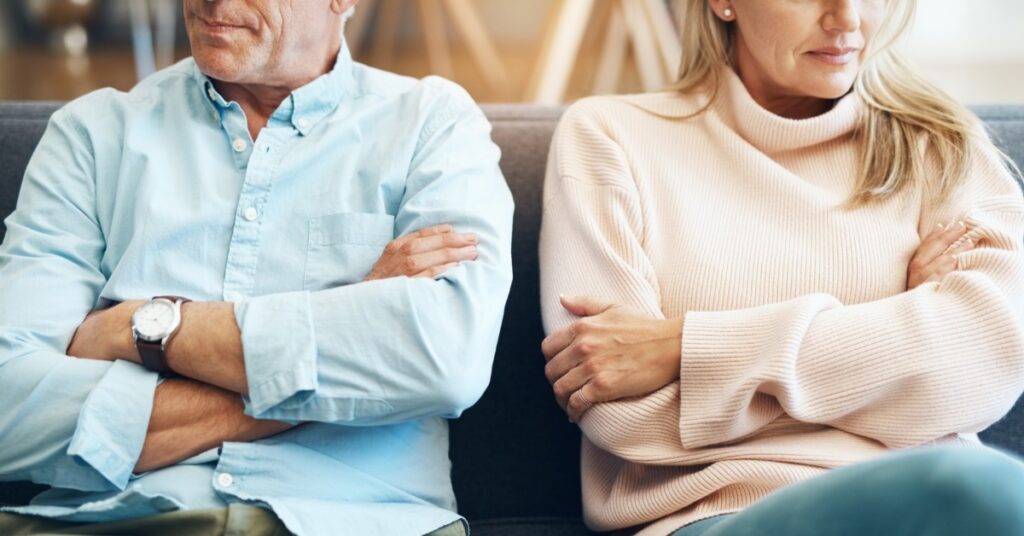 A man and woman sit on a couch, each with their arms crossed. They face forward while looking away from each other.