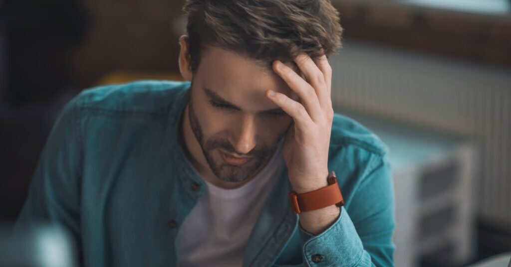 A young man sits at a desk and touches the left side of his face. He looks downward and has a neutral expression.