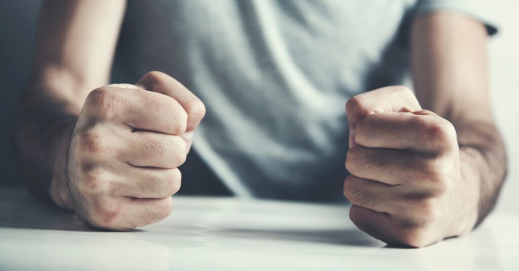 A person balls up their hands into tight fists and presses them down on a white table. They wear a light gray T-shirt.