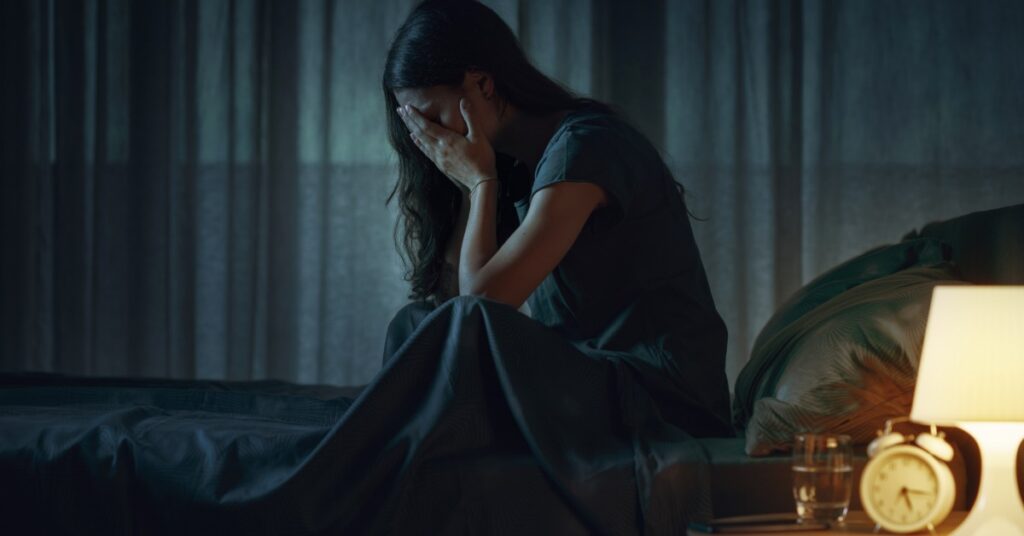 A woman sits up in bed with her hands covering her face. A lit lamp and an alarm clock are on the bedside table.