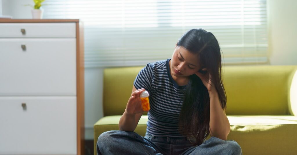 A woman sits cross-legged on the floor, holding a small orange pill bottle. She tilts her head and looks at the bottle.