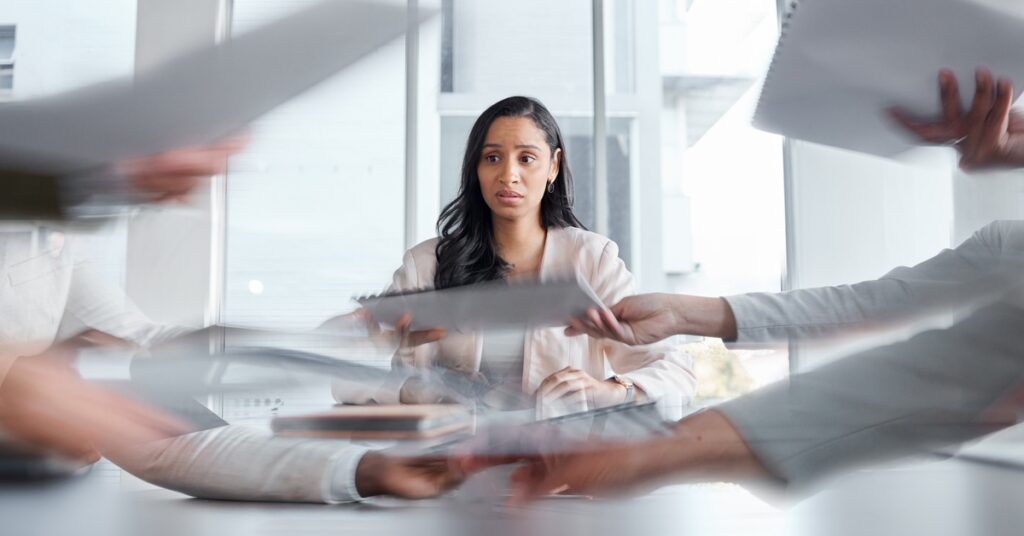 A woman sits at a conference table while several people hand papers across the table in a blur. She gazes elsewhere.