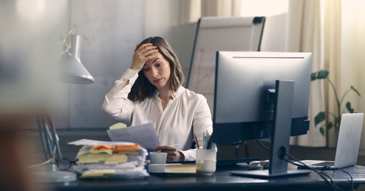 A woman presses her hand to her forehead, staring wide-eyed at a document. A thick stack of papers is on her desk.