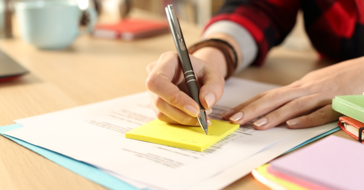 A person writes on yellow sticky notes placed over printed pages. Notebooks and other sticky notepads are nearby.
