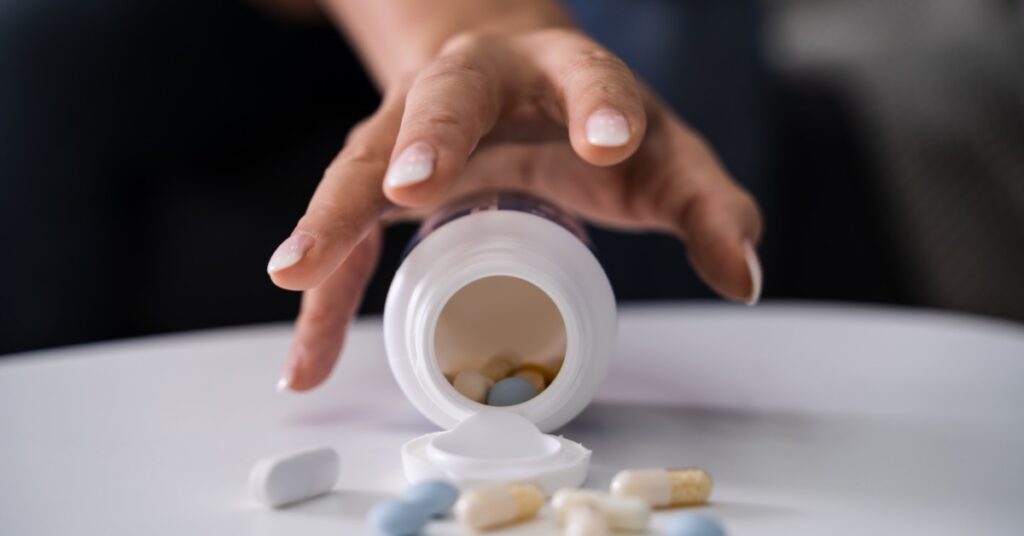 Someone reaching for an open pill bottle that is lying on a white table. Pills in different sizes and colors are on the table.