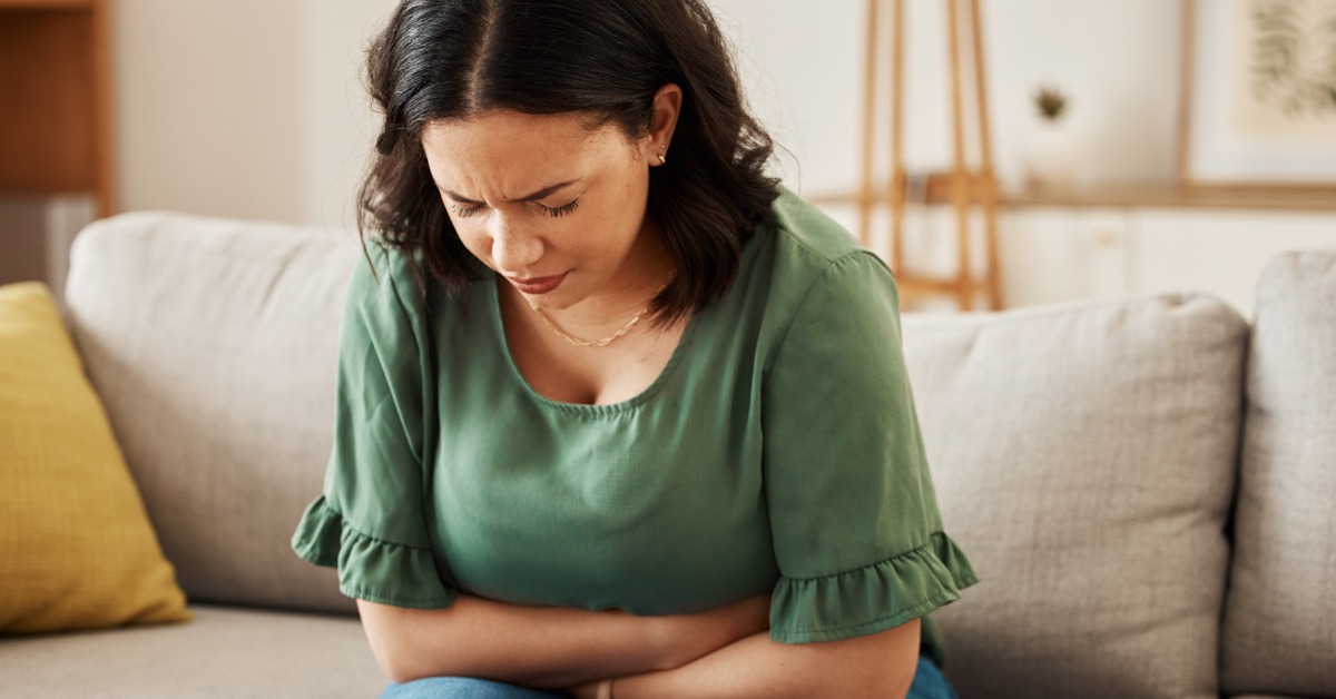 A woman sits on the edge of a couch with her head bent down, eyes closed, and arms crossed tightly over her stomach.