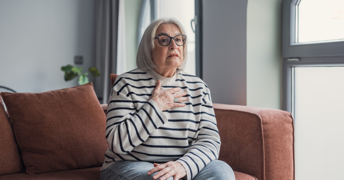 An older woman sits on the edge of a rust-colored couch. She rests one hand one her chest and looks forward.