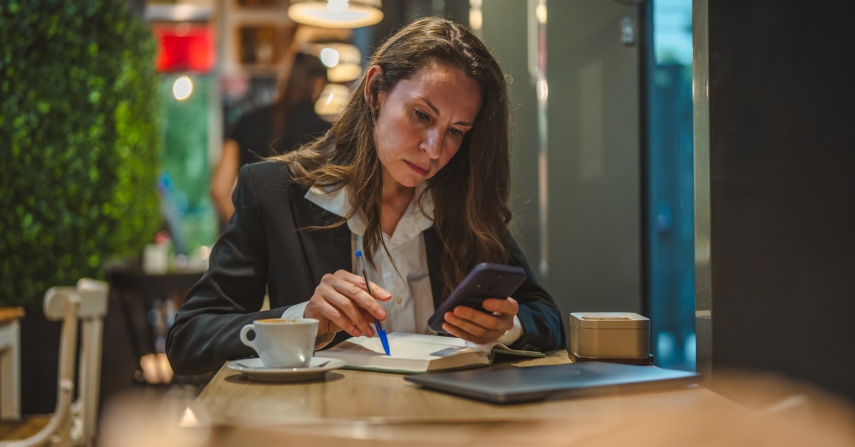 A woman at a cafe table looks at her smartphone. A coffee mug, a closed laptop, and a notebook are in front of her.