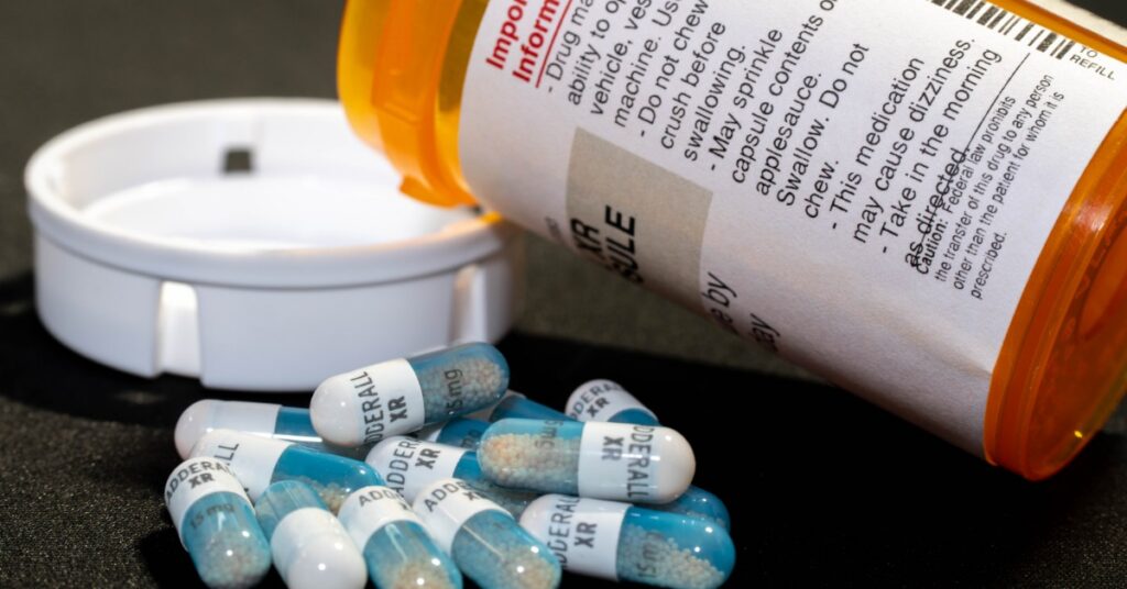Several blue-and-white Adderall XR capsules are scattered beside an open orange prescription bottle lying on its side.
