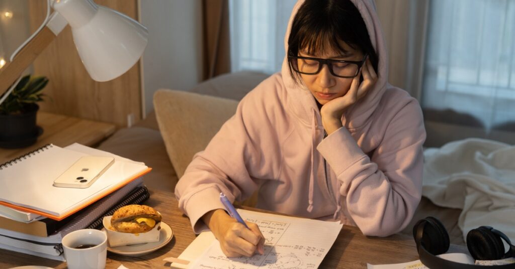 A woman rests her head on one hand while writing notes with the other. Study materials are spread out over the table.