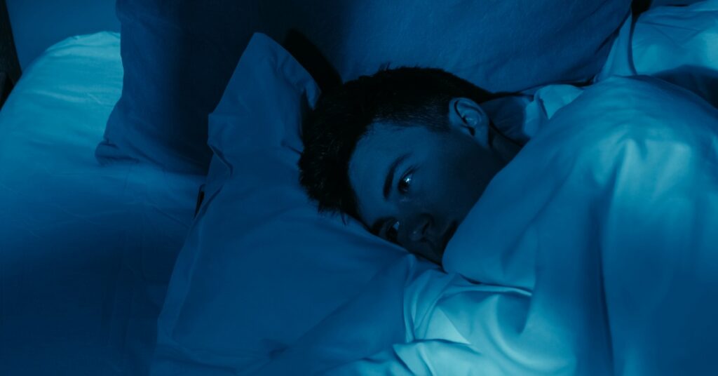 A man lies on his side with his eyes open beneath white bedding in a dark bedroom. Blue light falls across the bed.