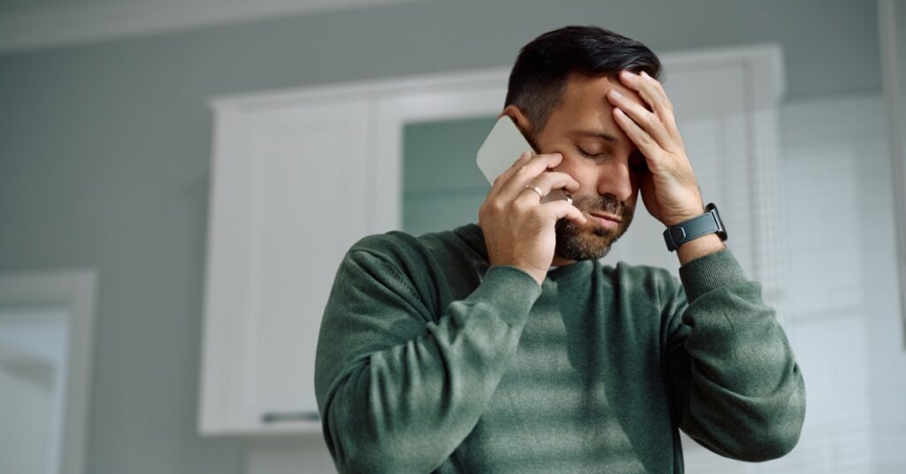 A standing man holds a smartphone to his ear. His eyes are closed, and he presses one hand against his forehead.