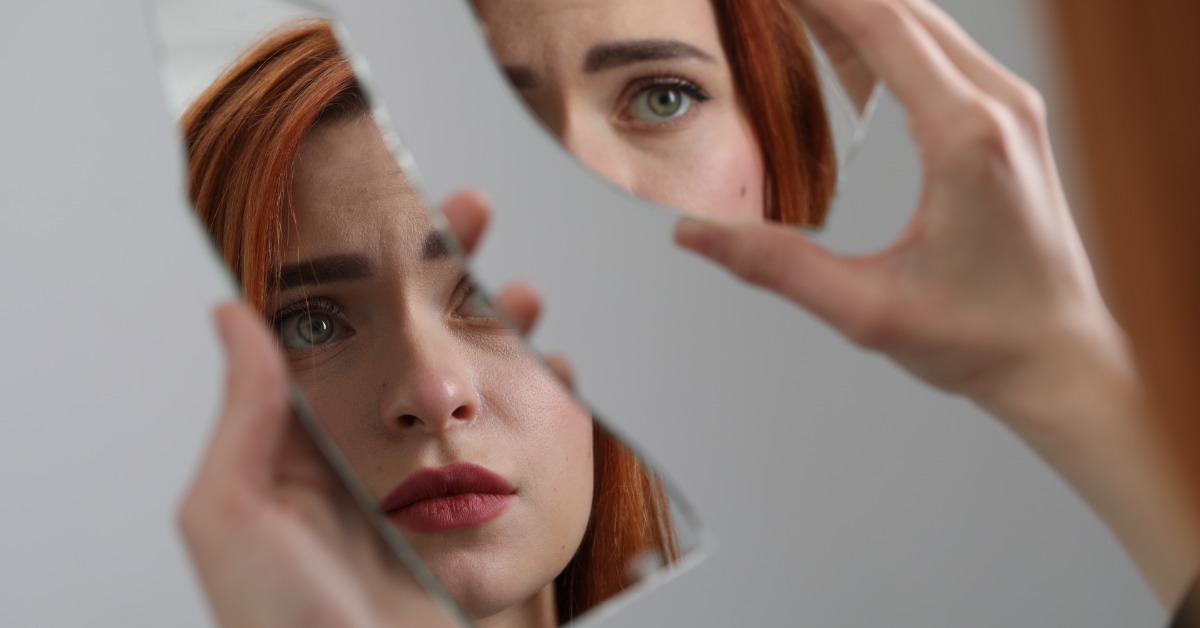 A red-haired woman holds two broken pieces of a mirror in front of her face. She looks at the shattered reflection.