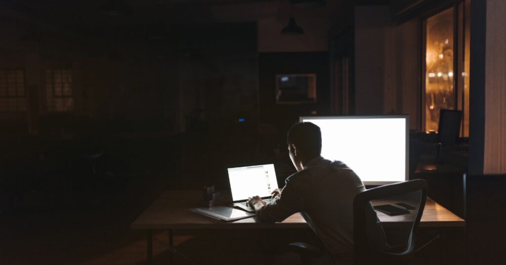 In a very dark room, a person is silhouetted by two bright computer screens. He leans forward and types on the laptop.