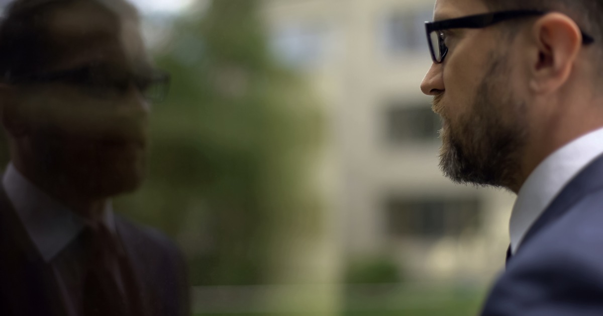 A bearded man in glasses and a suit stands facing a reflective glass surface. His reflection is dark and blurred.