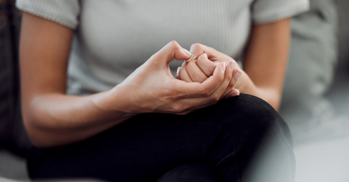 A woman sits with her legs crossed and her hands clasped tightly. She presses one thumb firmly with the other.