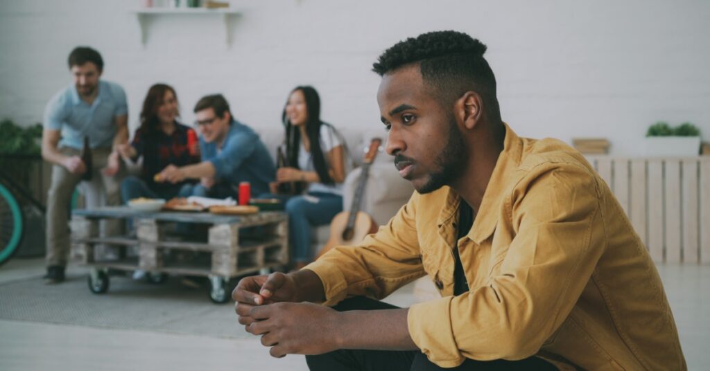 A man sits alone and gazes to the side. Behind him, several people sit on a couch, eating and talking together.